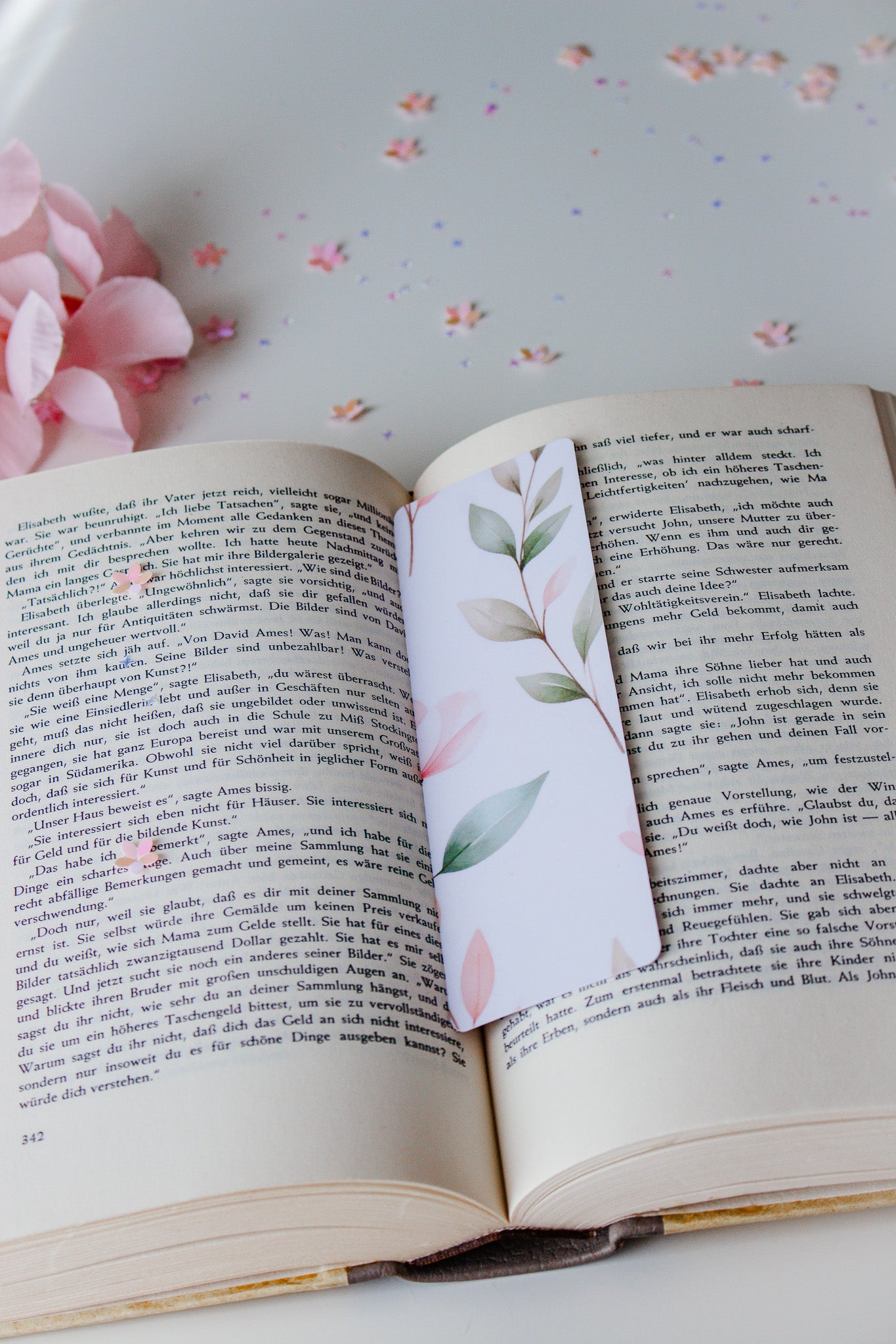 Aquarell-Lesezeichen mit grünen und rosafarbenen Blättern in einem aufgeschlagenen Buch, umgeben von buntem Konfetti und rosa Blütenblättern
