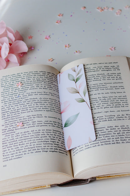 Aquarell-Lesezeichen mit grünen und rosafarbenen Blättern in einem aufgeschlagenen Buch, umgeben von buntem Konfetti und rosa Blütenblättern