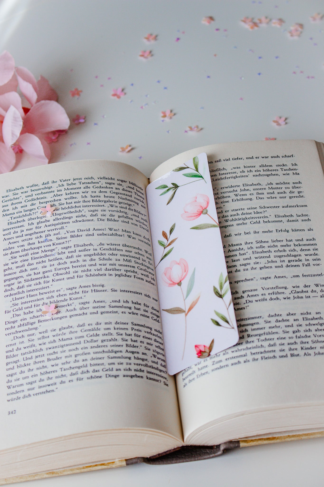 Aquarell-Lesezeichen mit rosa Tulpen und grünen Blättern in einem aufgeschlagenen Buch, umgeben von rosa Blütenblättern und buntem Konfetti
