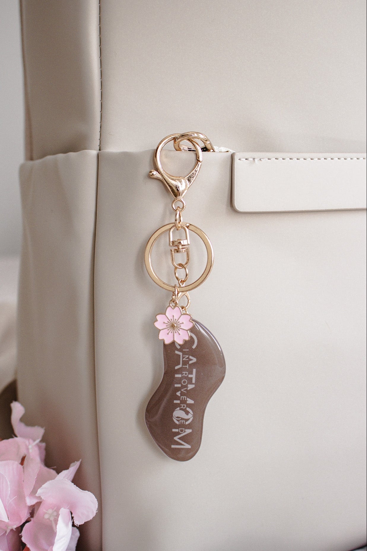 Brauner „Introverted Cat Mom" Schlüsselanhänger mit pinker Kirschblüte hängend an beiger Lederhandtasche, rosa Blüten im Hintergrund
