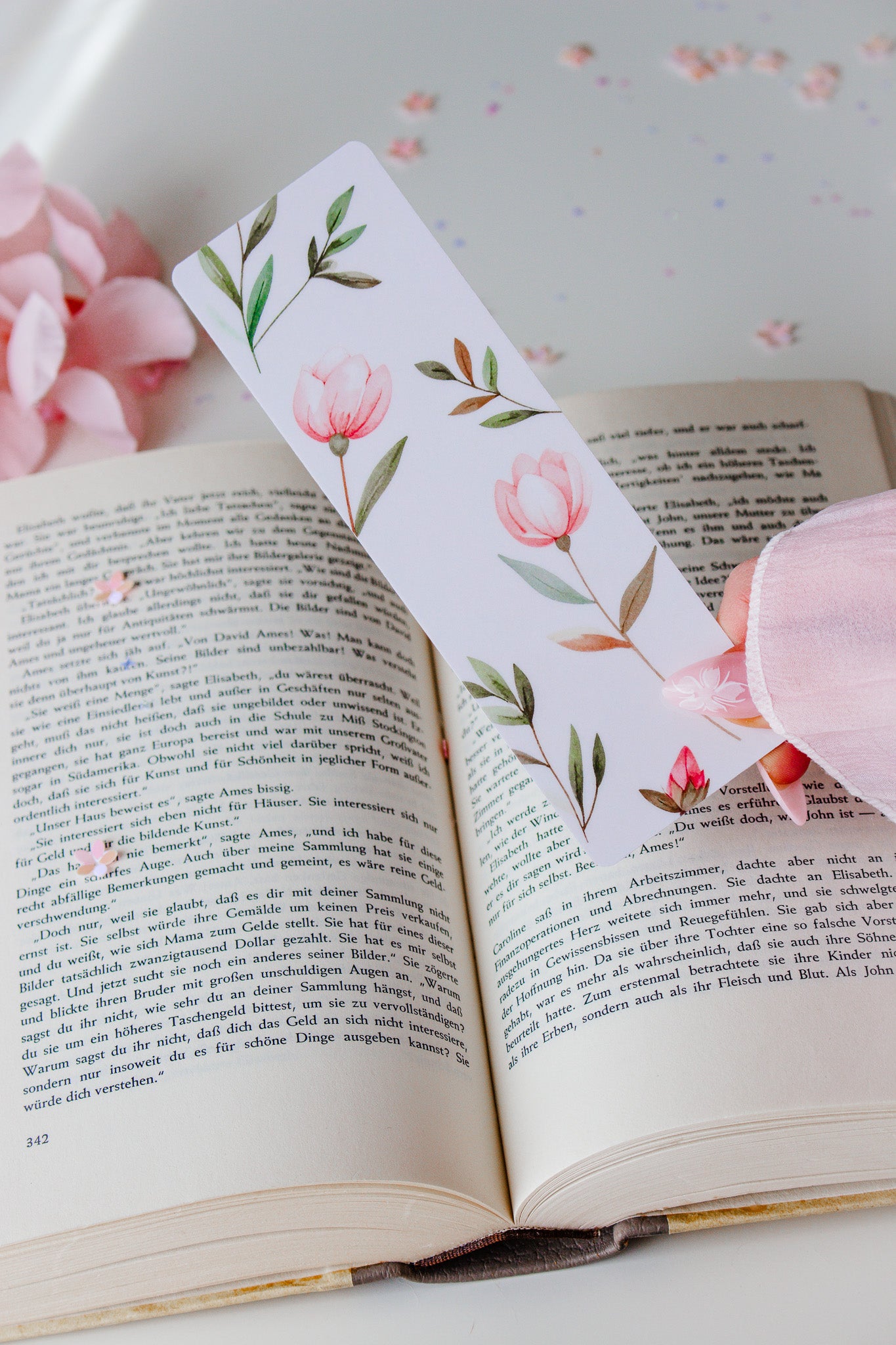 Doppelseitiges Lesezeichen mit Aquarell-Blumen-Motiv – rosa Tulpen und grüne Zweige auf weißem Hintergrund, gehalten über einem aufgeschlagenen Buch mit rosa Blütenblättern