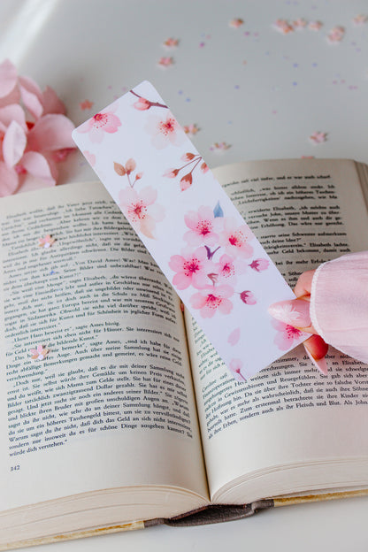 Doppelseitiges Lesezeichen mit Kirschblüten-Motiv in Rosa und Weiß, gehalten über einem aufgeschlagenen Buch vor hellem Hintergrund mit Blütenblättern