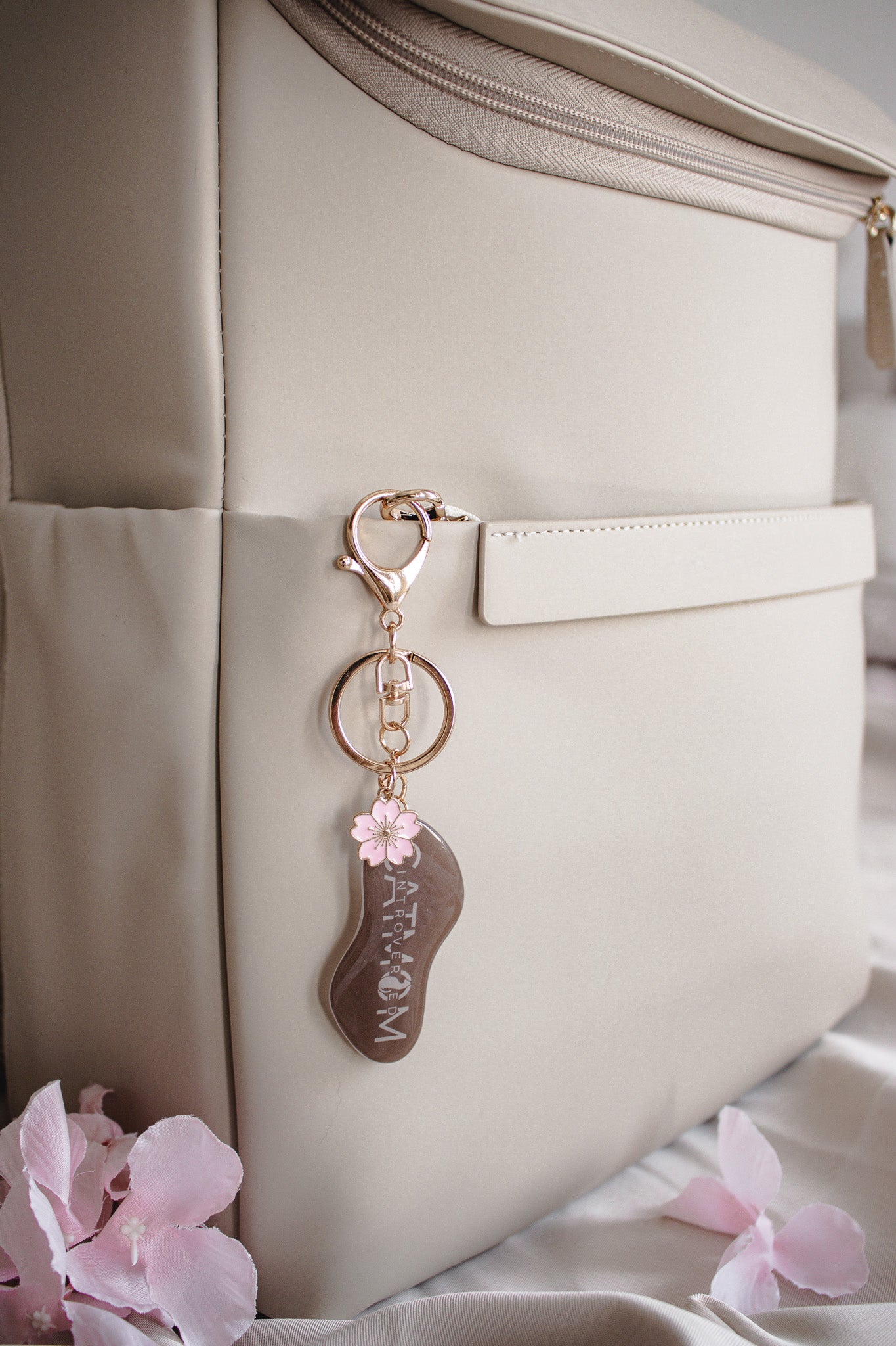 Handgemachter brauner Schlüsselanhänger „Introverted Cat Mom" mit Kirschblüten-Charm an beiger Handtasche, stimmungsvolle Flatlay-Aufnahme mit rosa Blüten
