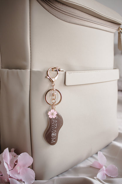 Handgemachter brauner Schlüsselanhänger „Introverted Cat Mom" mit Kirschblüten-Charm an beiger Handtasche, stimmungsvolle Flatlay-Aufnahme mit rosa Blüten
