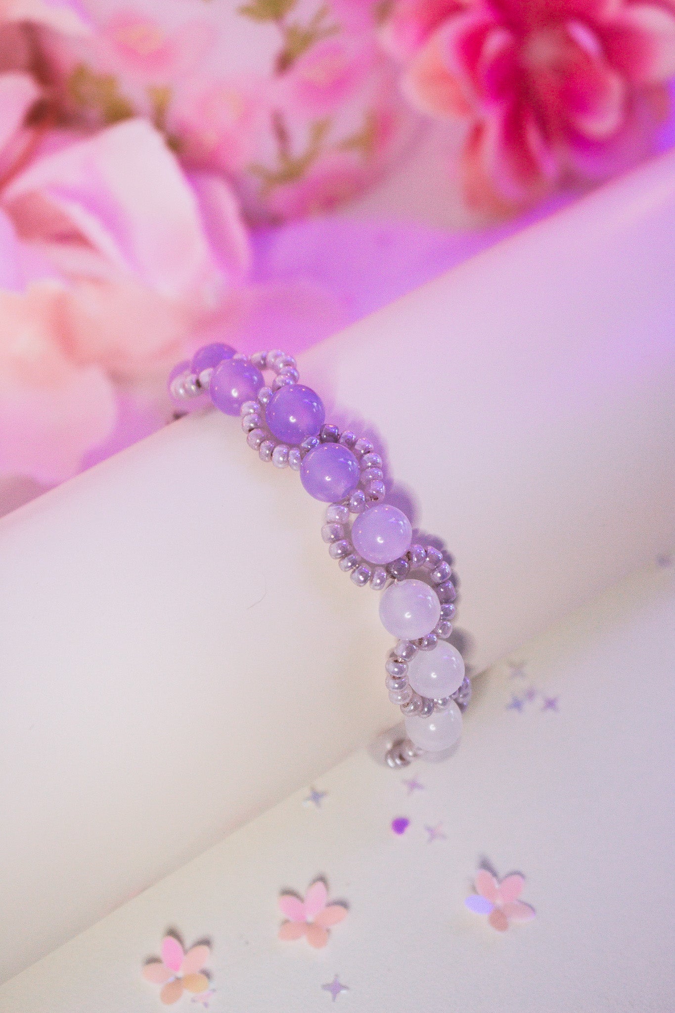 Perlenarmband mit lila und weißen Acrylperlen im Farbverlauf und Silberperlen, auf weißem Armband-Display vor rosa Blumendekoration