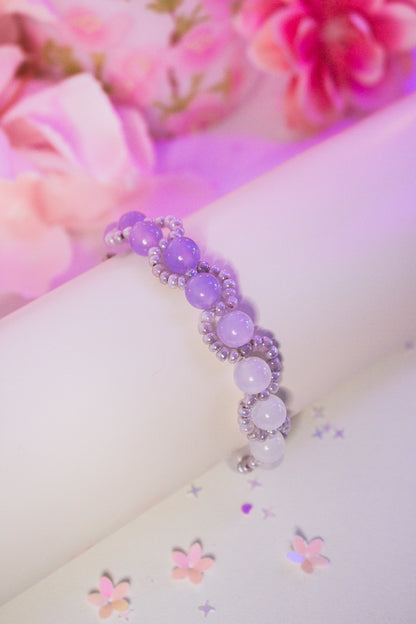 Perlenarmband mit lila und weißen Acrylperlen im Farbverlauf und Silberperlen, auf weißem Armband-Display vor rosa Blumendekoration
