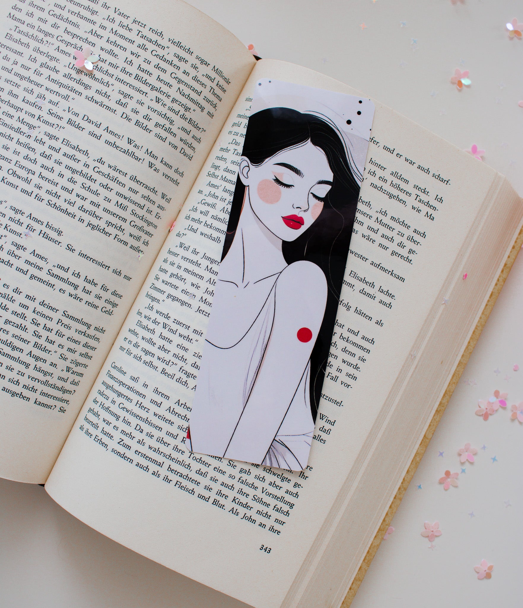 Laminiertes Lesezeichen mit minimalistischer Frauenillustration, schwarze Haare, rote Lippen und roter Akzentpunkt, schräg in aufgeschlagenem Buch mit Sternenkonfetti