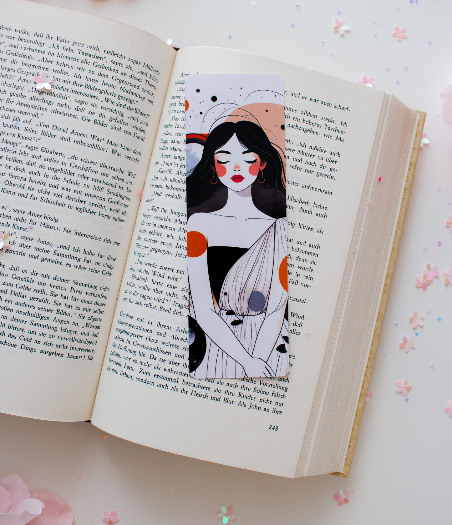 Laminiertes Lesezeichen mit abstrakter Celestial-Woman-Illustration – Frau mit roten Lippen, orangefarbenen und grauen Akzenten, aufgeschlagen im Buch mit Konfetti