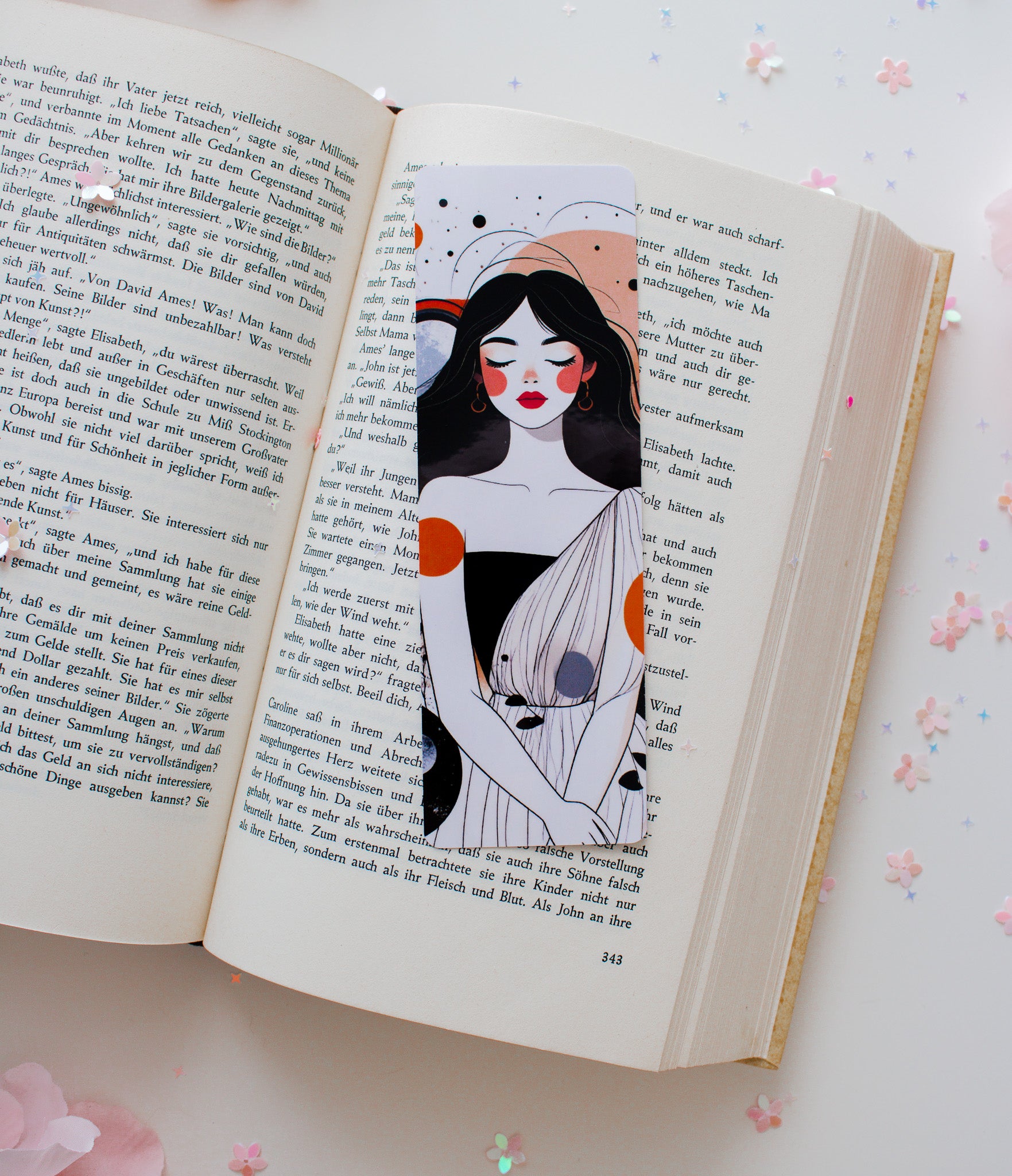 Laminiertes Lesezeichen mit abstrakter Celestial-Woman-Illustration – Frau mit roten Lippen, orangefarbenen und grauen Akzenten, aufgeschlagen im Buch mit Konfetti
