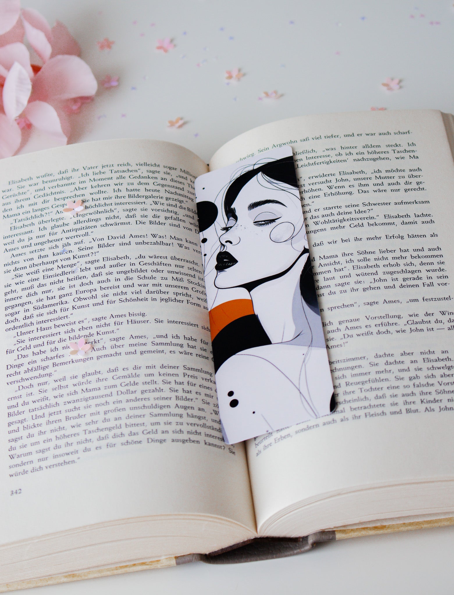 Laminiertes Lesezeichen mit minimalistischer Line-Art-Illustration einer Frau mit schwarzen Lippen und orangefarbenem Akzent, auf aufgeschlagenem Buch mit Konfetti