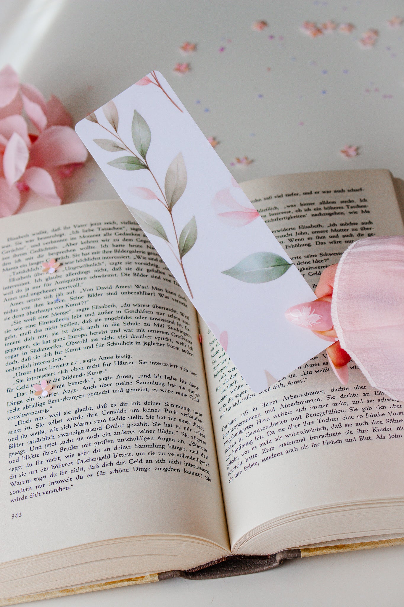 Doppelseitiges Lesezeichen mit Aquarell-Blätter-Motiv – zarte grüne und rosafarbene Blätter auf weißem Hintergrund, gehalten über einem aufgeschlagenen Buch mit rosa Blütenblättern