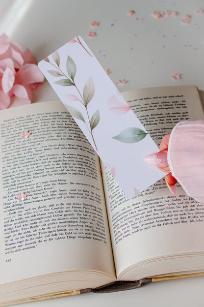 Doppelseitiges Lesezeichen mit Aquarell-Blätter-Motiv – zarte grüne und rosafarbene Blätter auf weißem Hintergrund, gehalten über einem aufgeschlagenen Buch mit rosa Blütenblättern