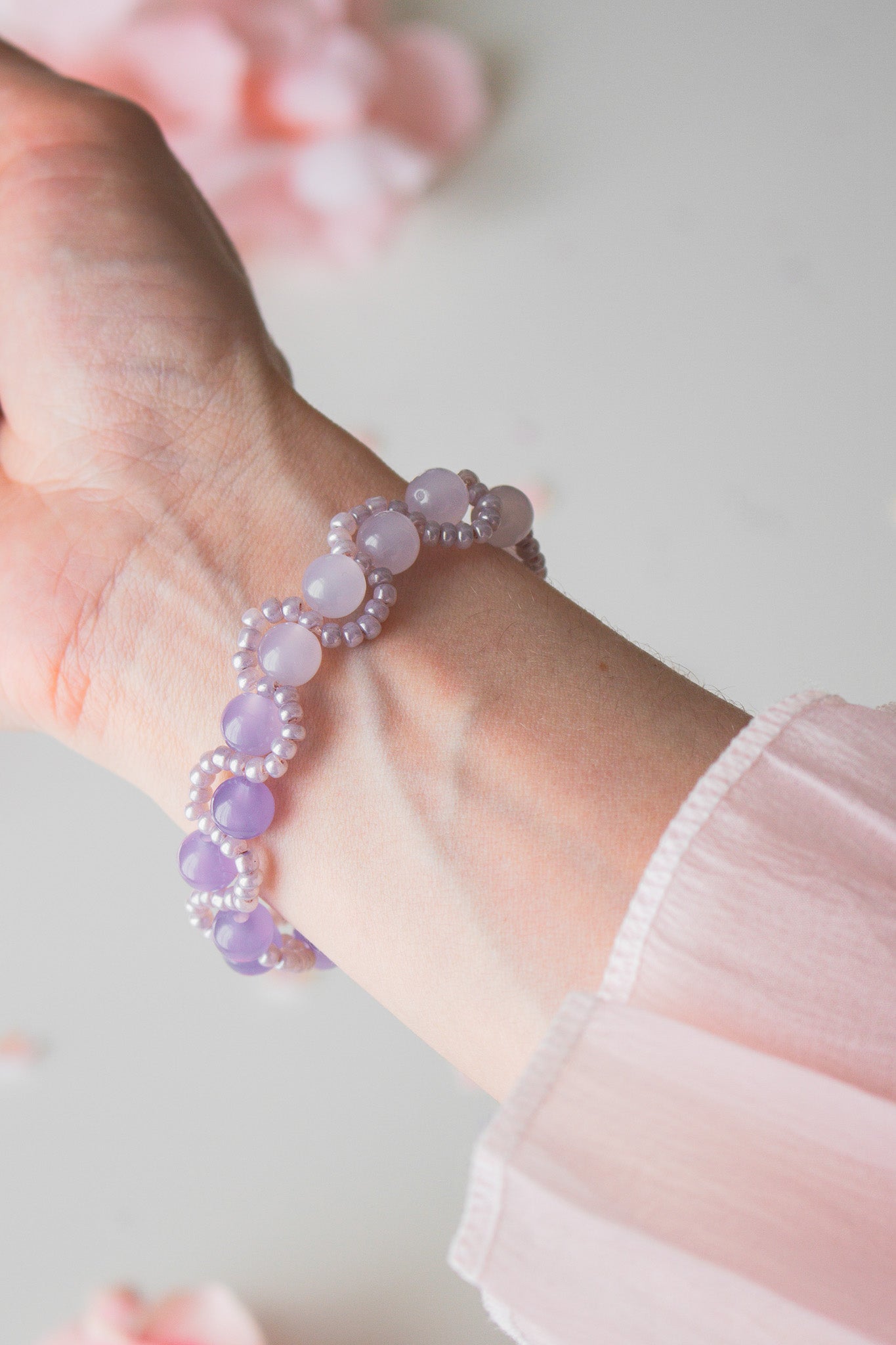 Zartes Perlenarmband mit helllila Acrylperlen und Silberperlen am Handgelenk, vor hellem Hintergrund mit Rosenblütenblättern