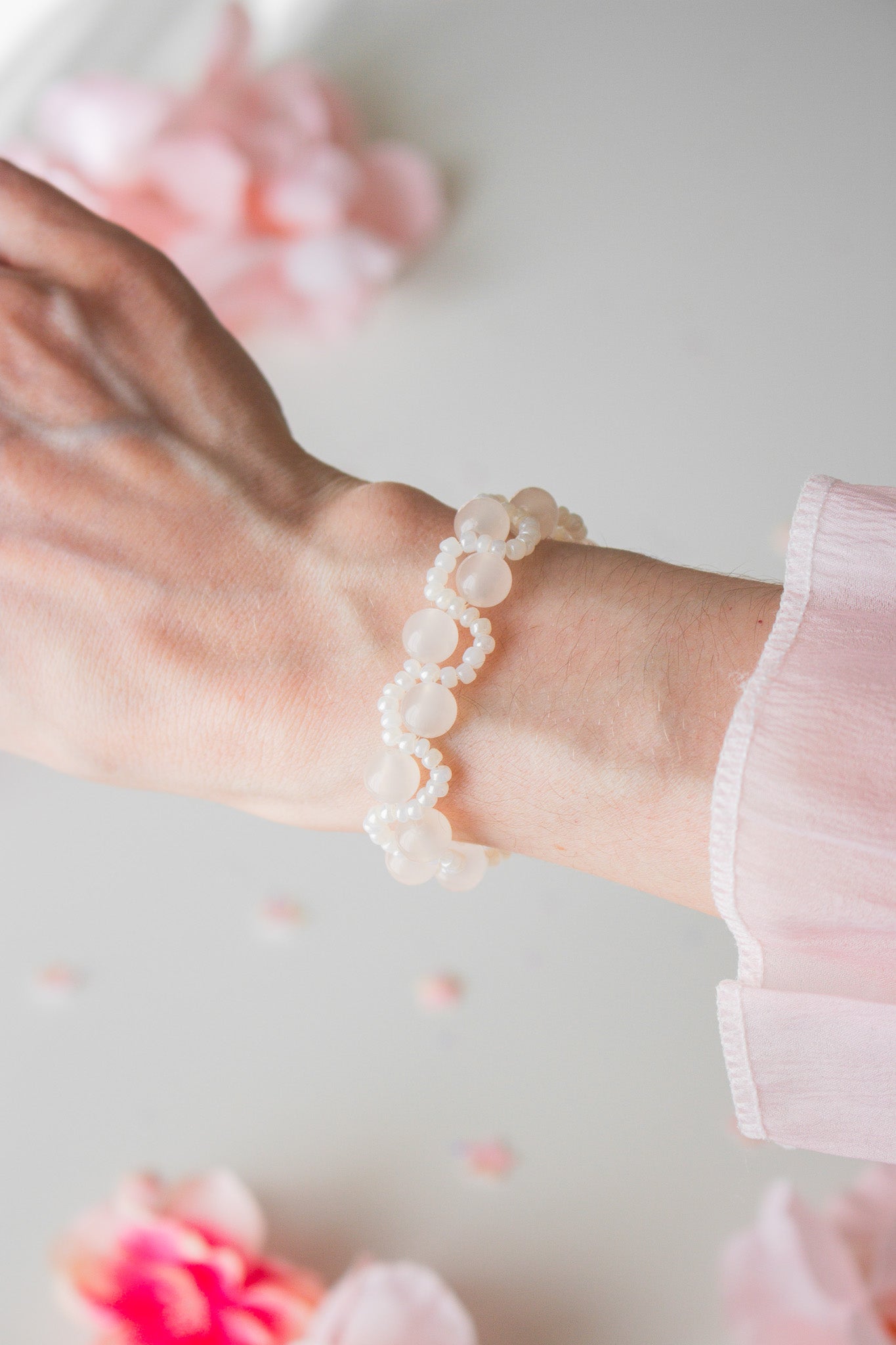 Elegantes Perlenarmband mit milchig-weißen Acrylperlen und weißen Rocailles am Handgelenk, vor hellem Hintergrund mit rosa Blütenblättern