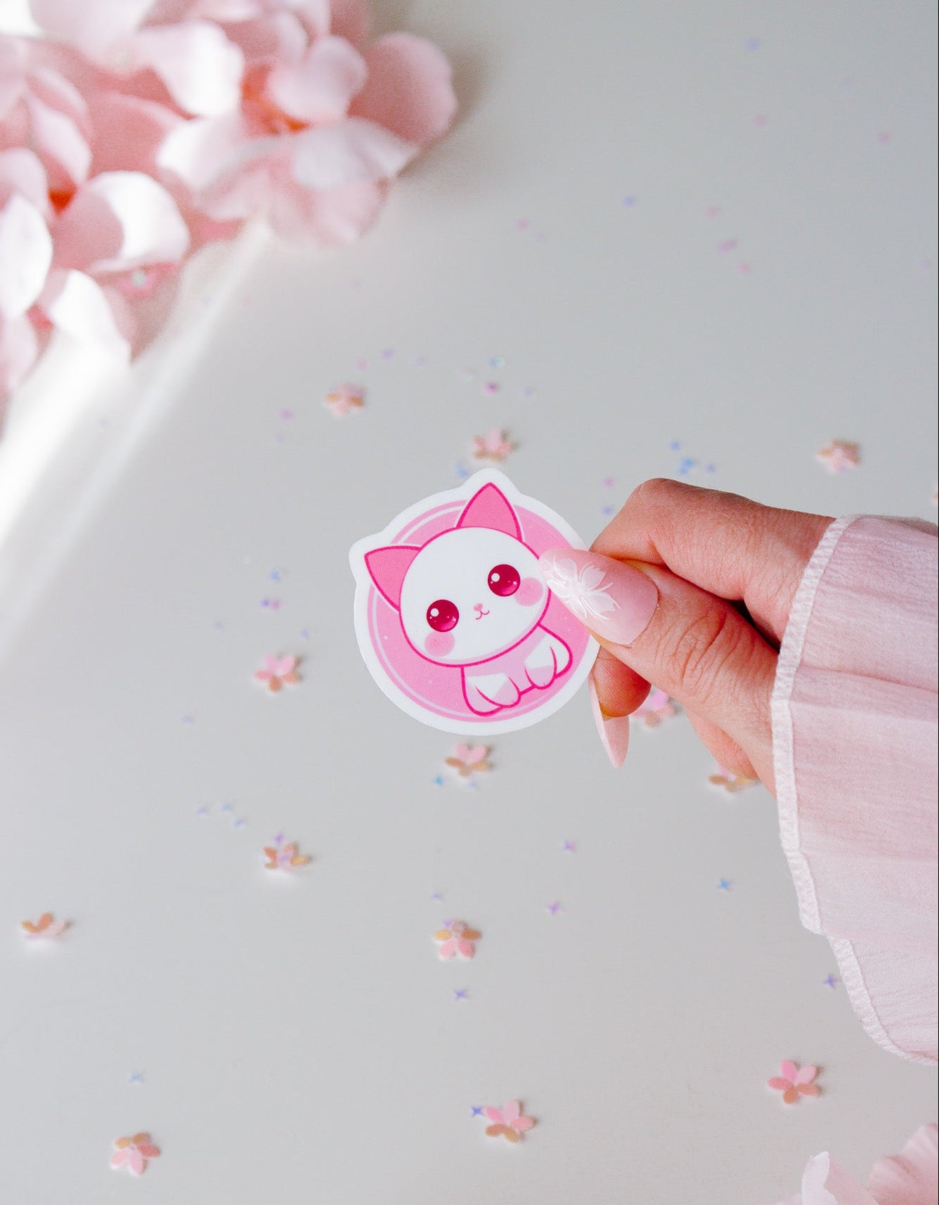 Kawaii Katzen-Sticker in Rosa und Weiß, gehalten von einer Hand vor weißem Hintergrund mit Rosenblütenblättern und kleinen Sternchen