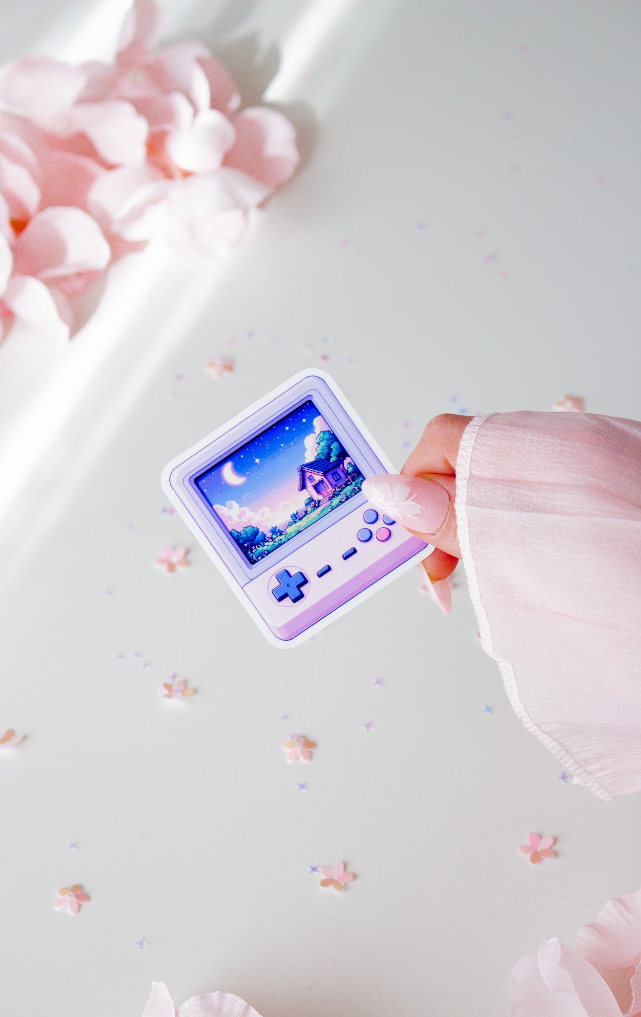 Niedlicher Retro-Konsolen-Sticker in Lila, Pixel-Art-Haus mit Mond und Sternen auf dem Display, in der Hand gehalten vor rosa Blüten und Sternenkonfetti