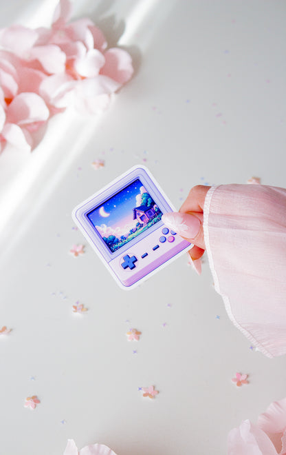 Niedlicher Retro-Konsolen-Sticker in Lila, Pixel-Art-Haus mit Mond und Sternen auf dem Display, in der Hand gehalten vor rosa Blüten und Sternenkonfetti