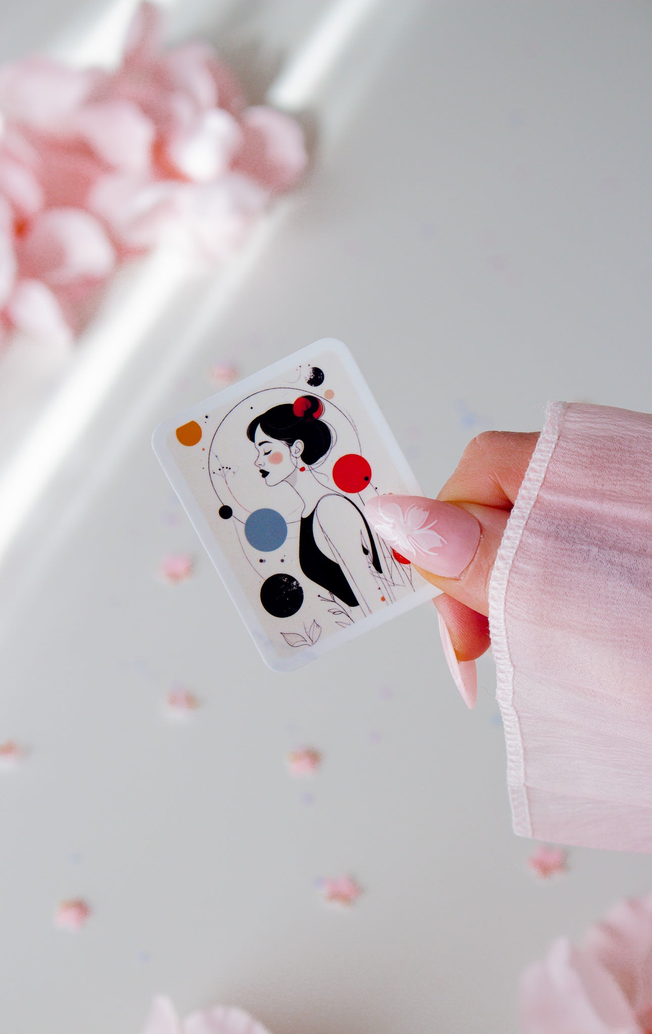 Vinyl Sticker mit minimalistischer Frauenillustration, schwarze Hochsteckfrisur, rote Ohrringe, abstrakte Kreise in Rot, Orange, Blau und Schwarz, in der Hand gehalten vor rosa Blüten
