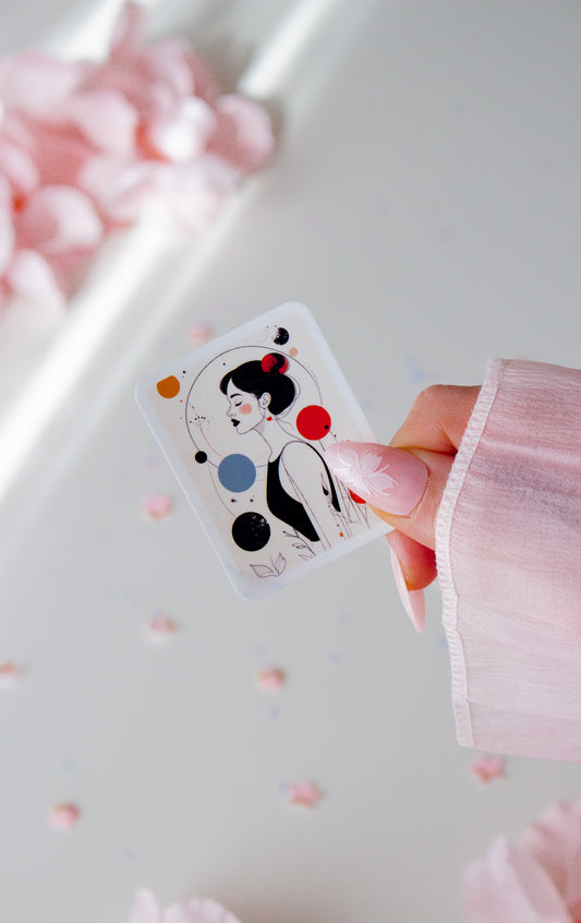 Vinyl Sticker mit minimalistischer Frauenillustration, schwarze Hochsteckfrisur, rote Ohrringe, abstrakte Kreise in Rot, Orange, Blau und Schwarz, in der Hand gehalten vor rosa Blüten
