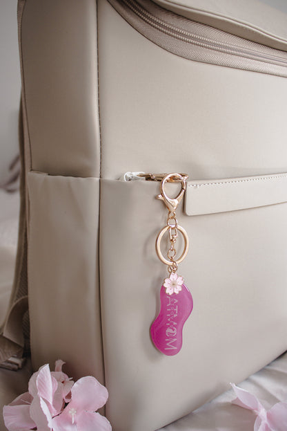 Pinker „Cat Mom" Schlüsselanhänger mit Kirschblüten-Detail und goldfarbenem Karabiner, hängend an einer beigen Tasche mit rosa Blüten im Hintergrund
