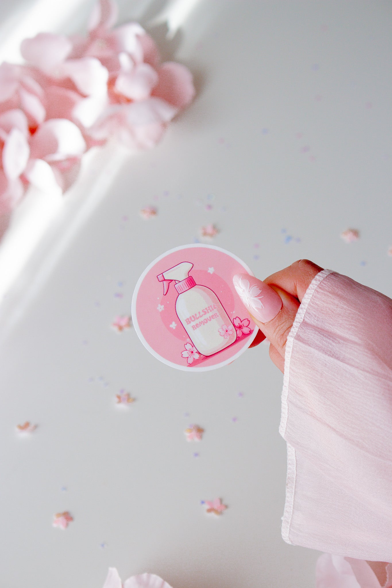 Kawaii Cleaning Spray Vinyl Sticker, runde Form mit pinker Sprühflasche und Sakura-Blüten auf rosa Hintergrund, in der Hand gehalten vor rosa Rosenblättern
