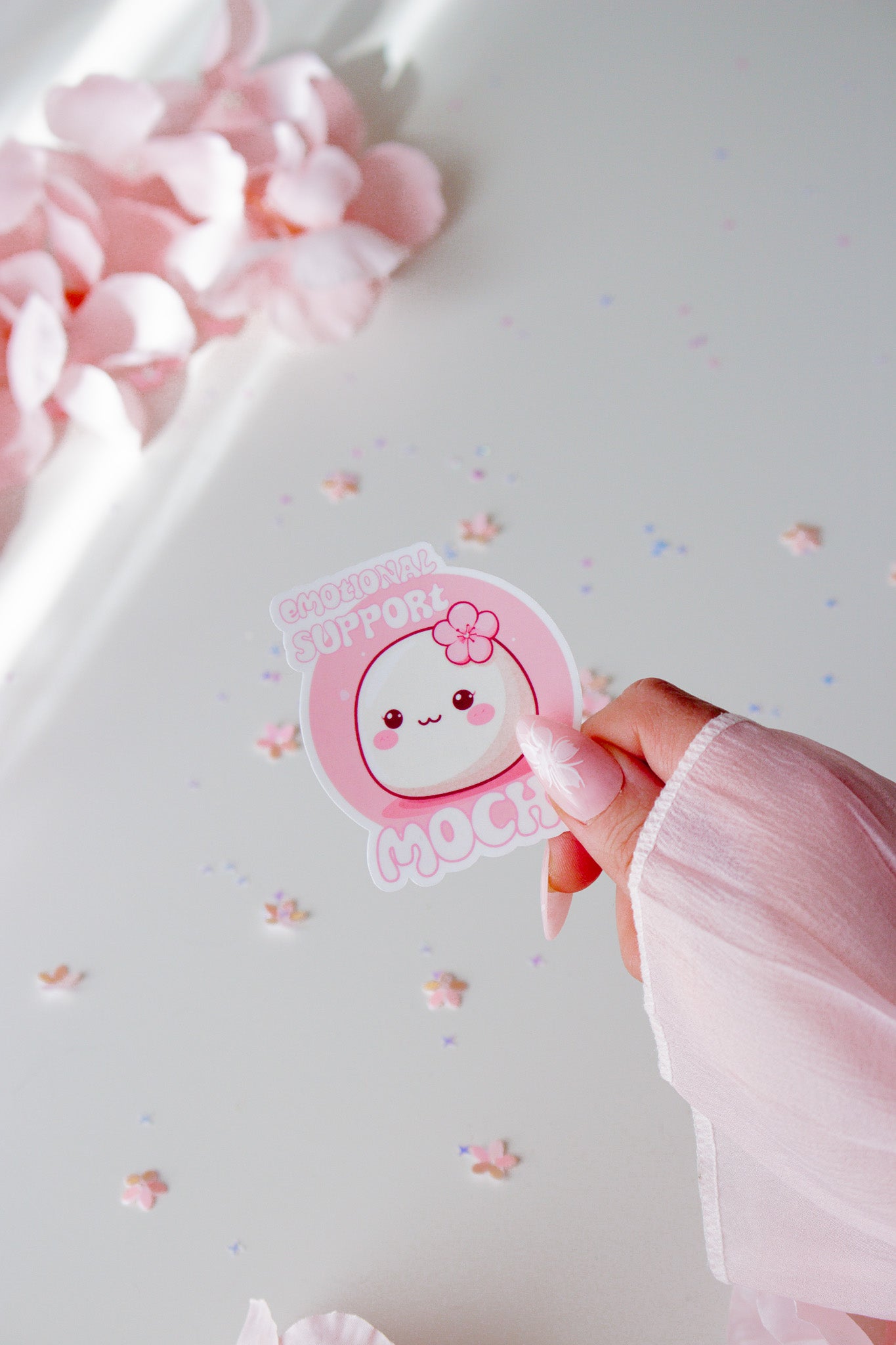 Kawaii Emotional Support Mochi Sticker, rundes weißes Mochi mit pinker Kirschblüte und lächelndem Gesicht auf rosa Hintergrund, in der Hand gehalten vor Sternenkonfetti

