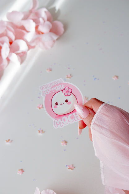 Kawaii Emotional Support Mochi Sticker, rundes weißes Mochi mit pinker Kirschblüte und lächelndem Gesicht auf rosa Hintergrund, in der Hand gehalten vor Sternenkonfetti

