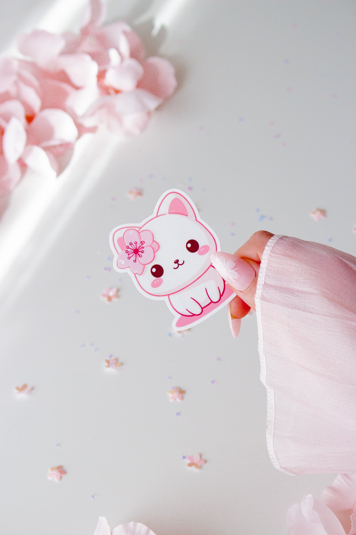 Kawaii Sakura Cat Vinyl Sticker, weißes Kätzchen mit pinker Kirschblüte, rosigen Wangen und großen Augen, in der Hand gehalten vor Sternenkonfetti