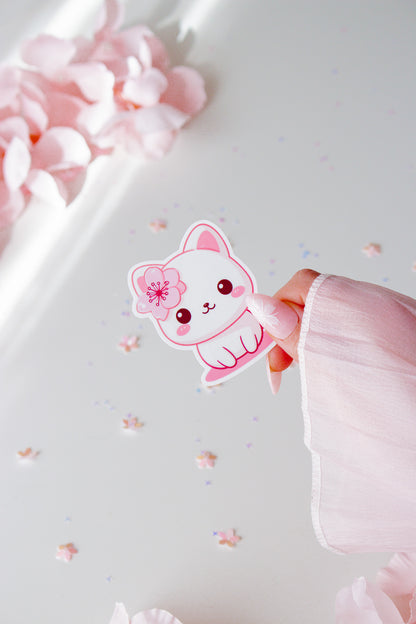 Kawaii Sakura Cat Vinyl Sticker, weißes Kätzchen mit pinker Kirschblüte, rosigen Wangen und großen Augen, in der Hand gehalten vor Sternenkonfetti