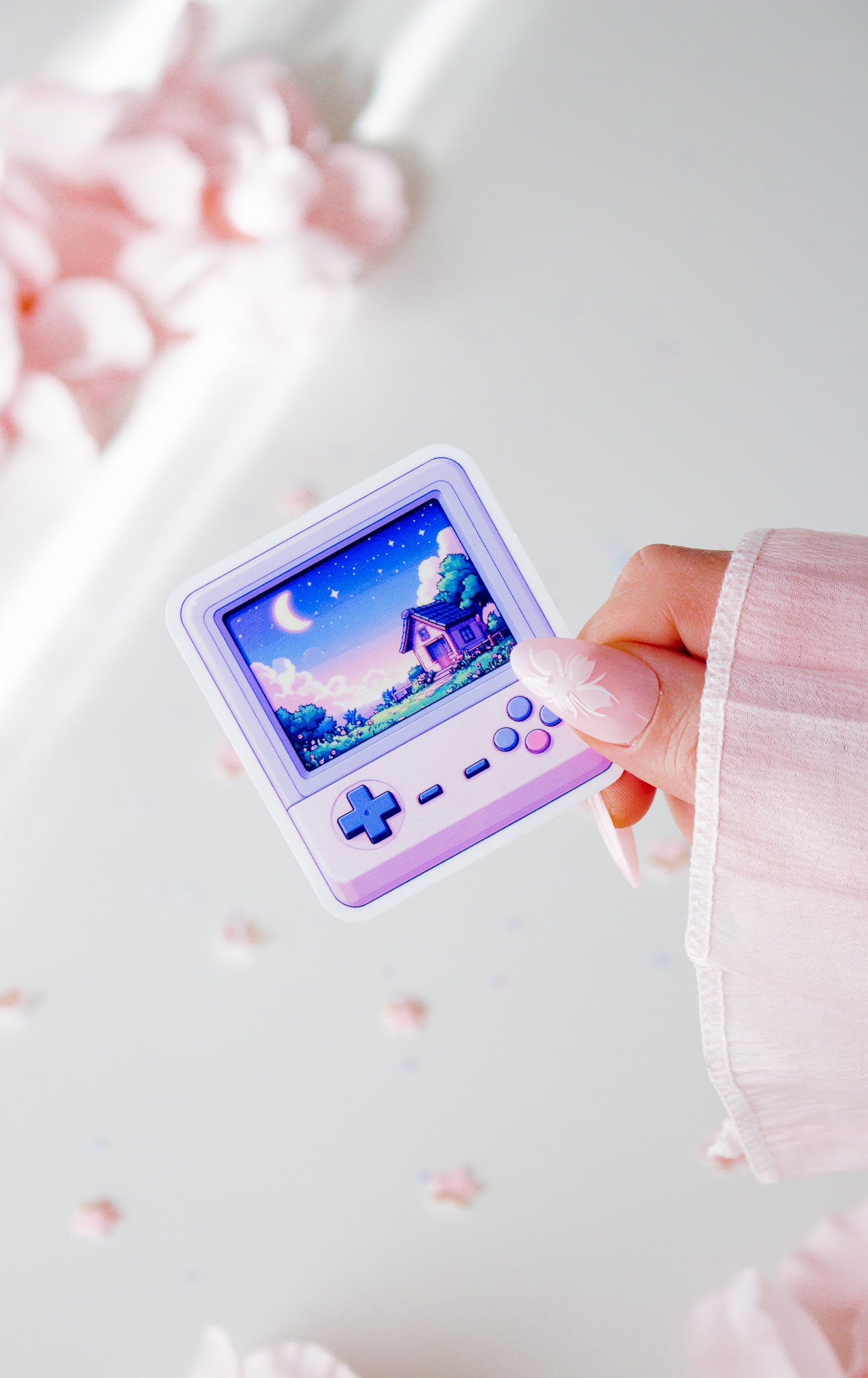 Kawaii Vinyl Sticker, lila Retro-Handheld-Konsole mit Pixel-Art-Cottage und Mondnacht-Szene, in der Hand gehalten vor rosa Blüten