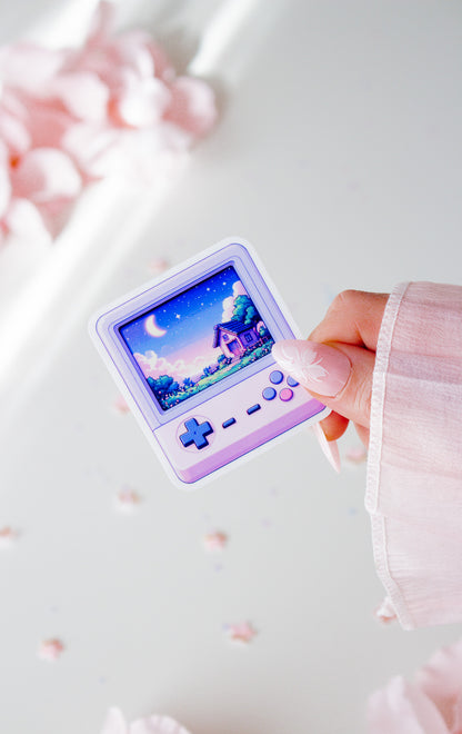 Kawaii Vinyl Sticker, lila Retro-Handheld-Konsole mit Pixel-Art-Cottage und Mondnacht-Szene, in der Hand gehalten vor rosa Blüten