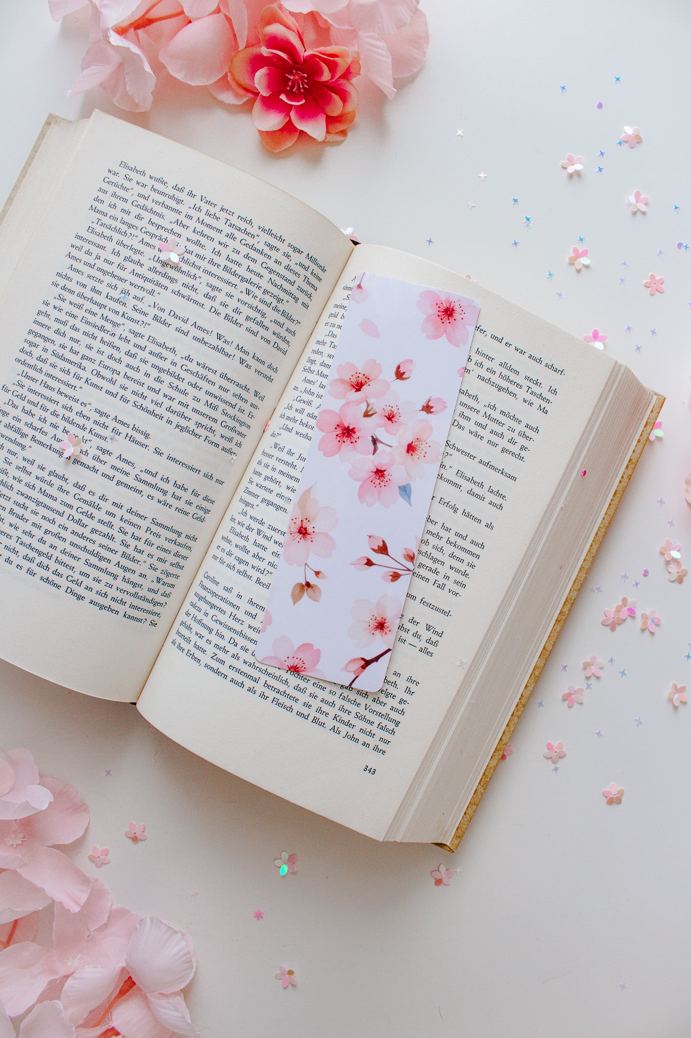 Kirschblüten-Lesezeichen in Rosa und Weiß in einem aufgeschlagenen Buch, umgeben von rosa Blüten und Glitzerkonfetti
