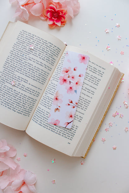 Kirschblüten-Lesezeichen in Rosa und Weiß in einem aufgeschlagenen Buch, umgeben von rosa Blüten und Glitzerkonfetti