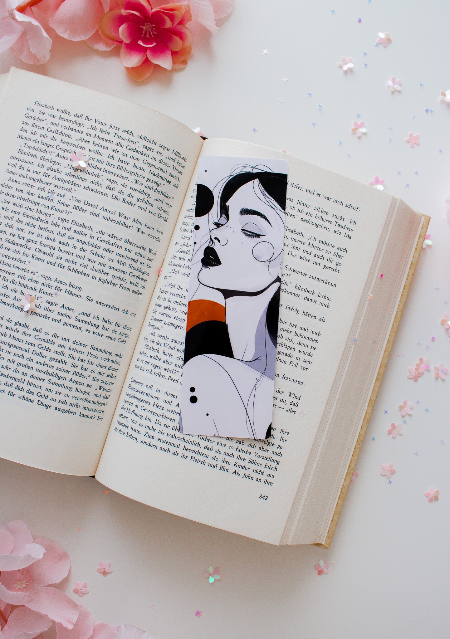 Laminiertes Glossy-Lesezeichen mit abstrakter Frauenillustration, schwarze Haare, orangefarbener Streifen, auf aufgeschlagenem Buch mit rosa Blüten

