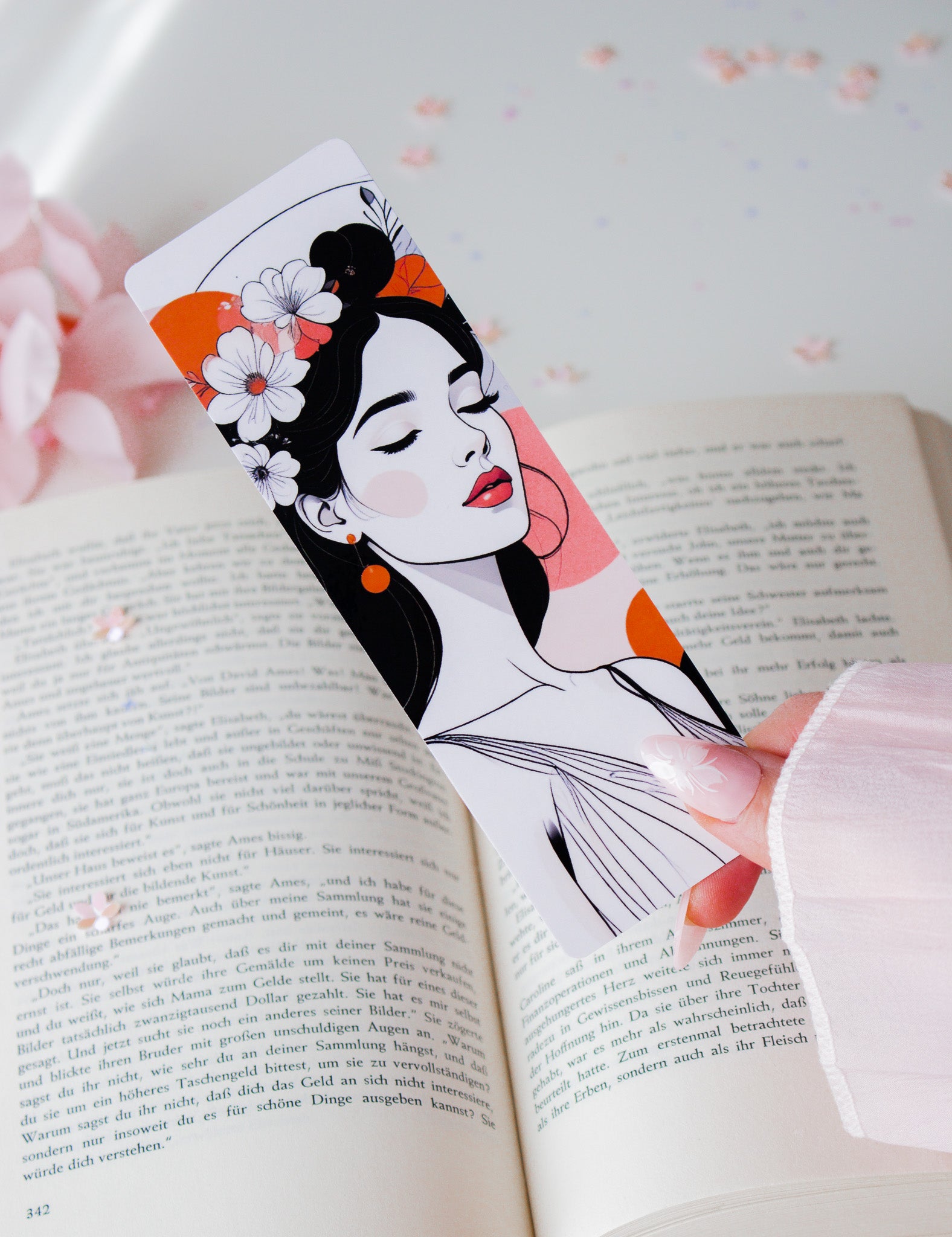 Laminiertes Lesezeichen mit Floral-Woman-Illustration in der Hand gehalten – weiße Blüten im schwarzen Haar, rote Lippen, orangefarbene Akzente, aufgeschlagenes Buch im Hintergrund