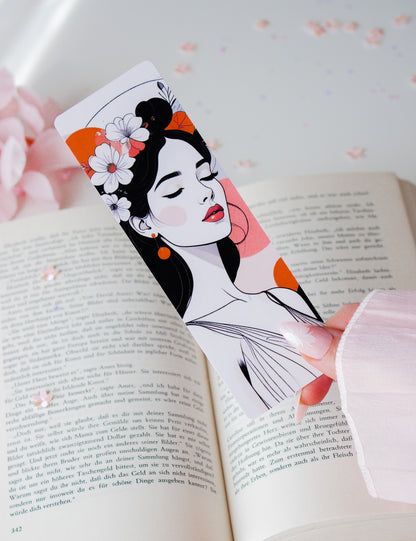 Laminiertes Lesezeichen mit Floral-Woman-Illustration in der Hand gehalten – weiße Blüten im schwarzen Haar, rote Lippen, orangefarbene Akzente, aufgeschlagenes Buch im Hintergrund