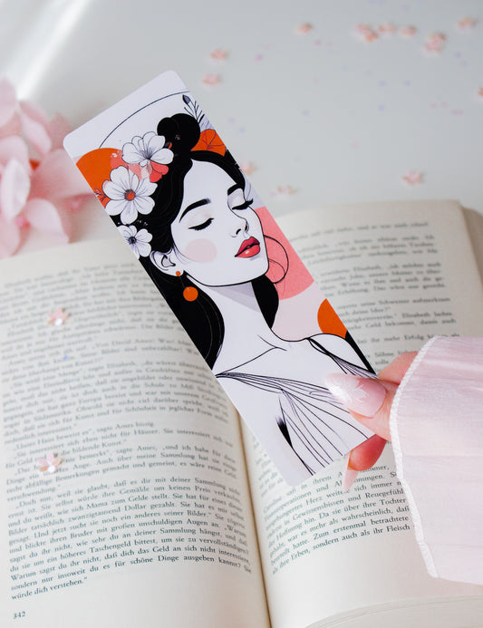 Laminiertes Lesezeichen mit Floral-Woman-Illustration in der Hand gehalten – weiße Blüten im schwarzen Haar, rote Lippen, orangefarbene Akzente, aufgeschlagenes Buch im Hintergrund