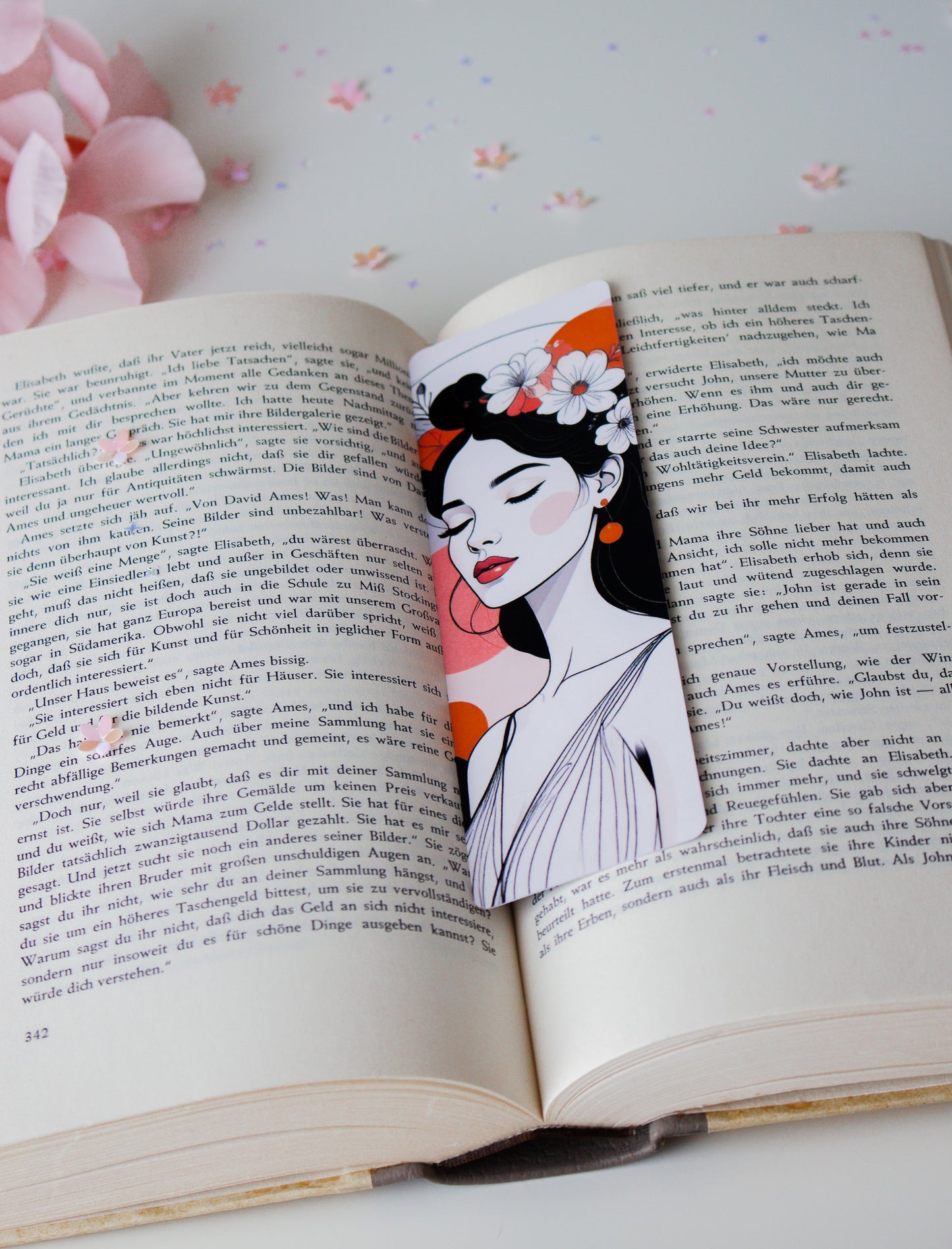 Laminiertes Lesezeichen mit eleganter Frauenillustration, weiße Blüten im schwarzen Haar, rote Lippen und orangefarbene Akzente, schräg in aufgeschlagenem Buch