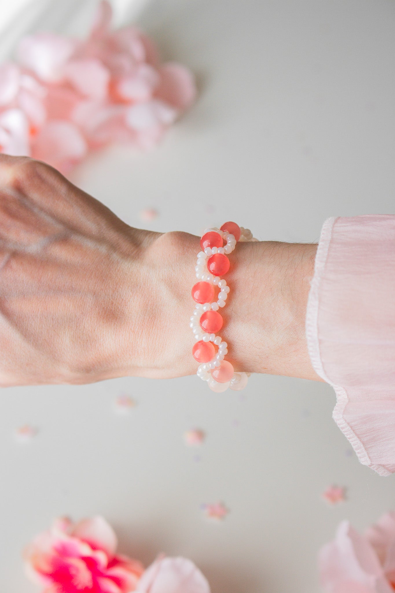 Perlenarmband mit korallenroten Acrylperlen und weißen Rocailles am Handgelenk, vor hellem Hintergrund mit rosa Blütenblättern