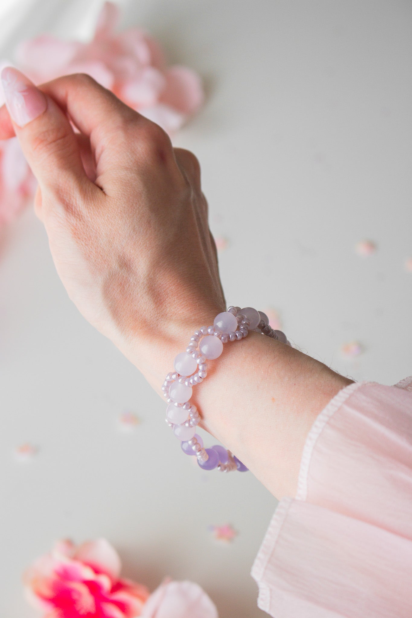 Perlenarmband mit lila Acrylperlen in verschiedenen Tönen und Silberperlen am Handgelenk, vor hellem Hintergrund mit Rosenblütenblättern