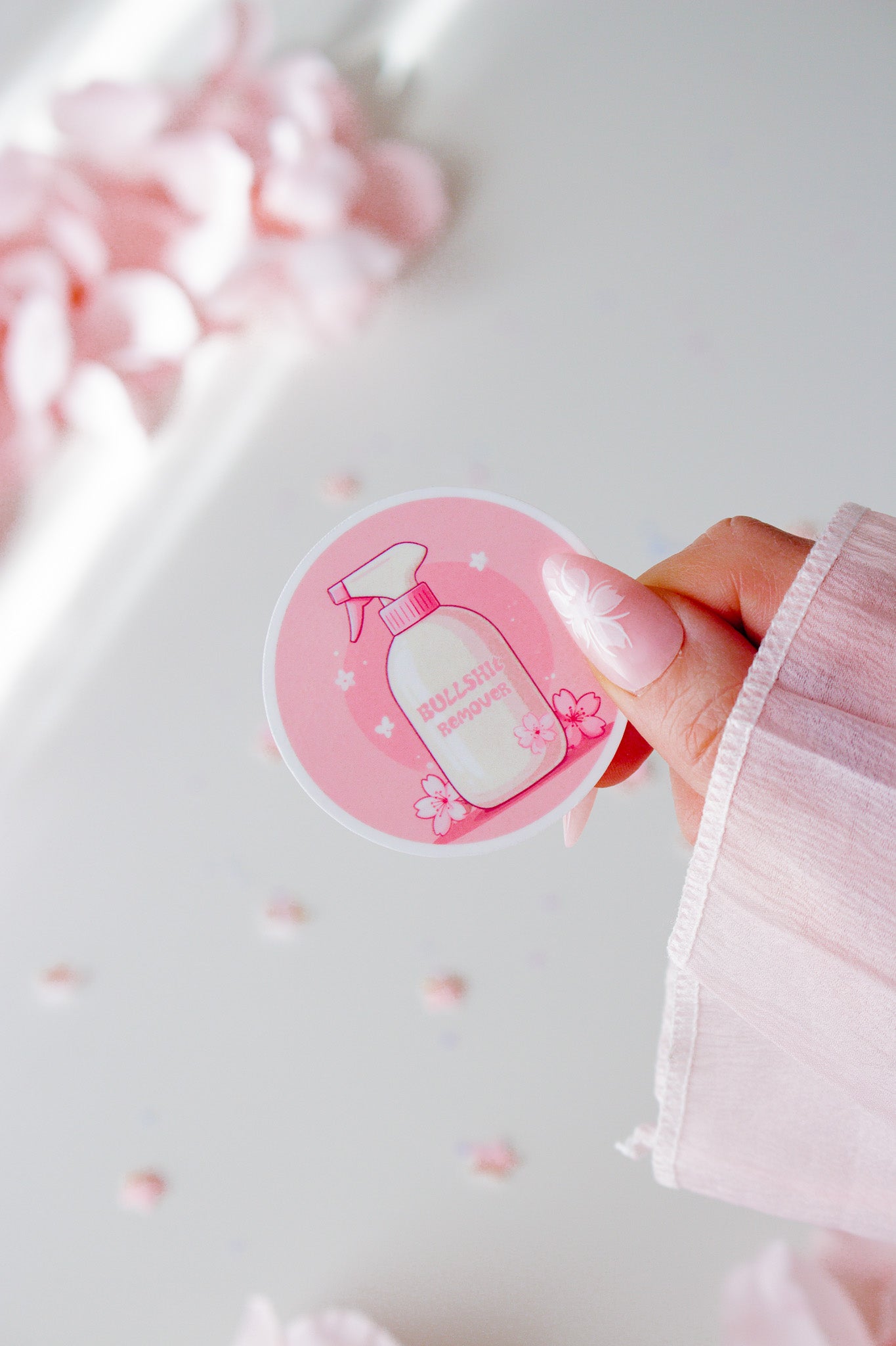 Runder Vinyl Sticker mit kawaii Sprühflasche in Rosa und Weiß, Kirschblüten-Dekoration auf rosa Hintergrund, in der Hand gehalten vor rosa Blüten