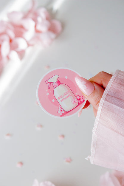 Runder Vinyl Sticker mit kawaii Sprühflasche in Rosa und Weiß, Kirschblüten-Dekoration auf rosa Hintergrund, in der Hand gehalten vor rosa Blüten