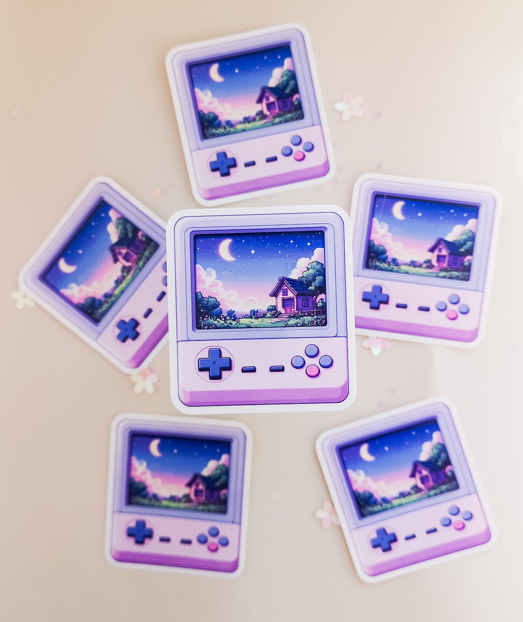 Sechs kawaii Retro-Handheld-Konsolen-Sticker in Lila mit Pixel-Art-Cottage und Mondnacht-Szene, Draufsicht auf beigem Hintergrund mit Sternenkonfetti

