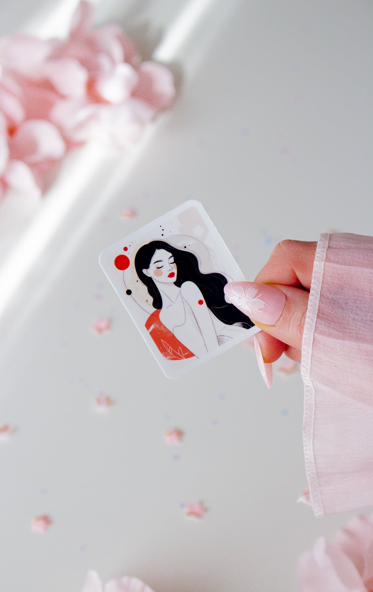 Vinyl Sticker mit minimalistischer Frauenillustration, schwarze lange Haare, rote Lippen und roter Kreis-Akzent, in der Hand gehalten vor rosa Blüten