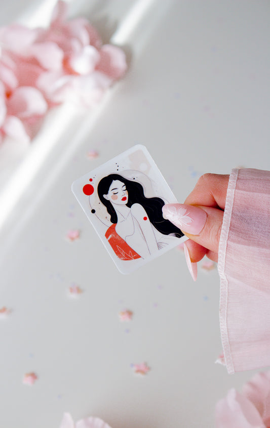 Vinyl Sticker mit minimalistischer Frauenillustration, schwarze lange Haare, rote Lippen und roter Kreis-Akzent, in der Hand gehalten vor rosa Blüten