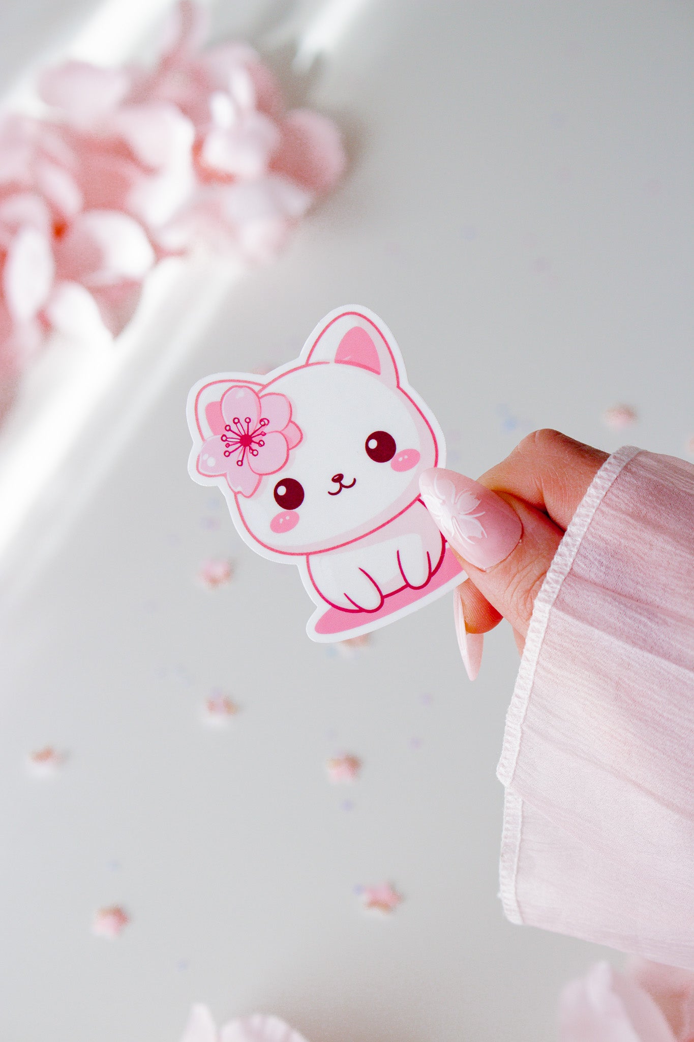 Vinyl Sticker mit niedlichem weißem Kätzchen und pinker Kirschblüte im Haar, rosige Wangen und große Augen, in der Hand gehalten vor rosa Blüten