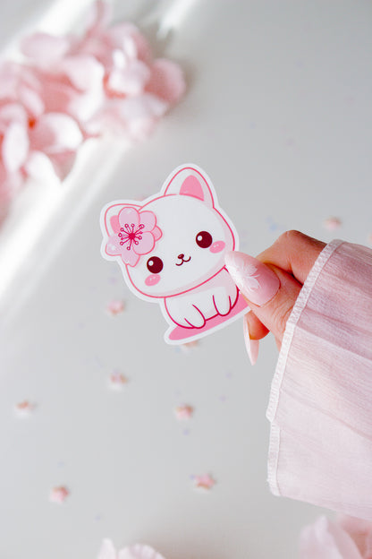 Vinyl Sticker mit niedlichem weißem Kätzchen und pinker Kirschblüte im Haar, rosige Wangen und große Augen, in der Hand gehalten vor rosa Blüten