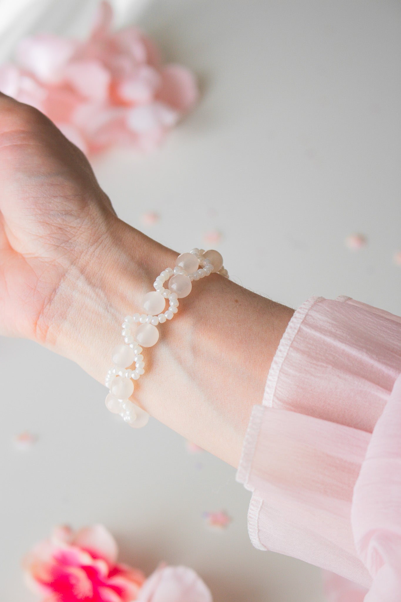 Zartes Perlenarmband mit cremefarbenen Acrylperlen und weißen Rocailles am Handgelenk, vor hellem Hintergrund mit rosa Blütenblättern