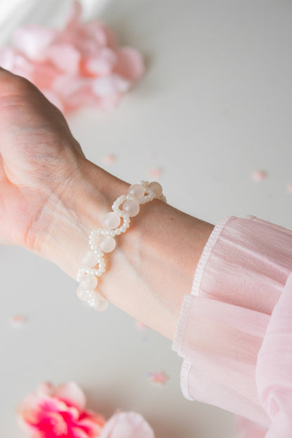 Zartes Perlenarmband mit cremefarbenen Acrylperlen und weißen Rocailles am Handgelenk, vor hellem Hintergrund mit rosa Blütenblättern